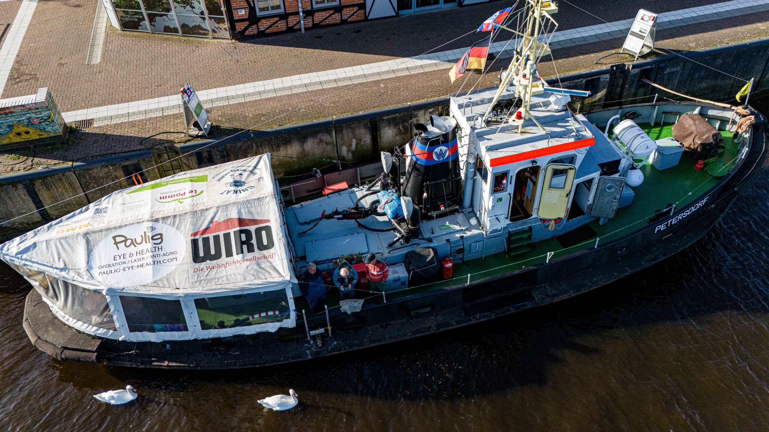 Ein älteres Boot mit Planendach auf einem Fluss; Schwäne schwimmen daneben; bunte Details.